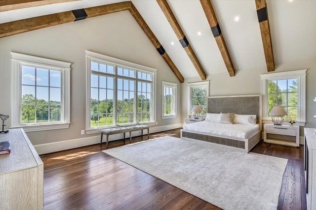 Bedroom with vaulted ceiling, exposed reclaimed wood beams, large windows, hardwood floors, and modern furniture in a bright, natural setting.