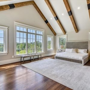 Bedroom with vaulted ceiling, exposed reclaimed wood beams, large windows, hardwood floors, and modern furniture in a bright, natural setting.